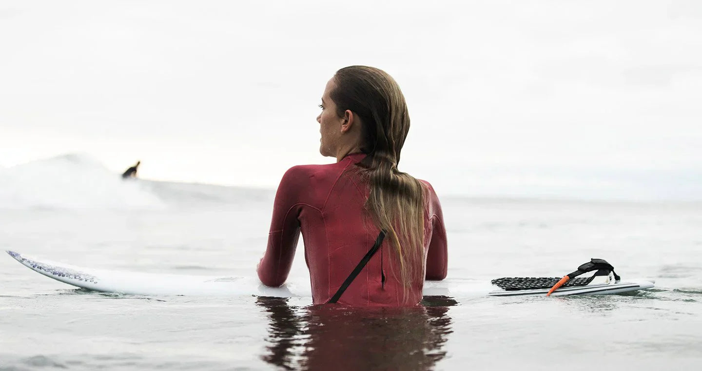 A female surfer in a red wetsuit sits on her surfboard in calm ocean waters, gazing towards the horizon. Another surfer rides a wave in the background.