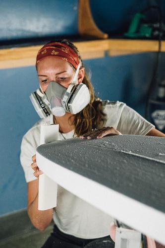Woman shaping a surfboard in a workshop, wearing a respirator and headband.