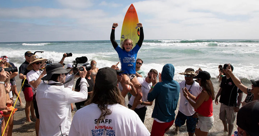 Samantha Sibley celebrates victory at the Super Girl Pro surfing competition, surrounded by cheering crowd on the beach.