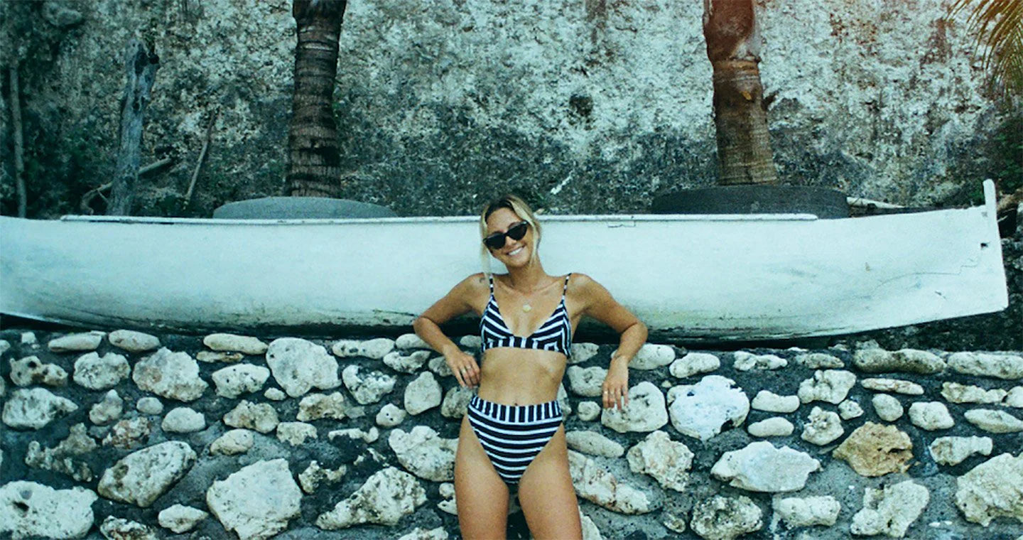 Woman in striped bikini posing by a stone wall and boat under palm trees.