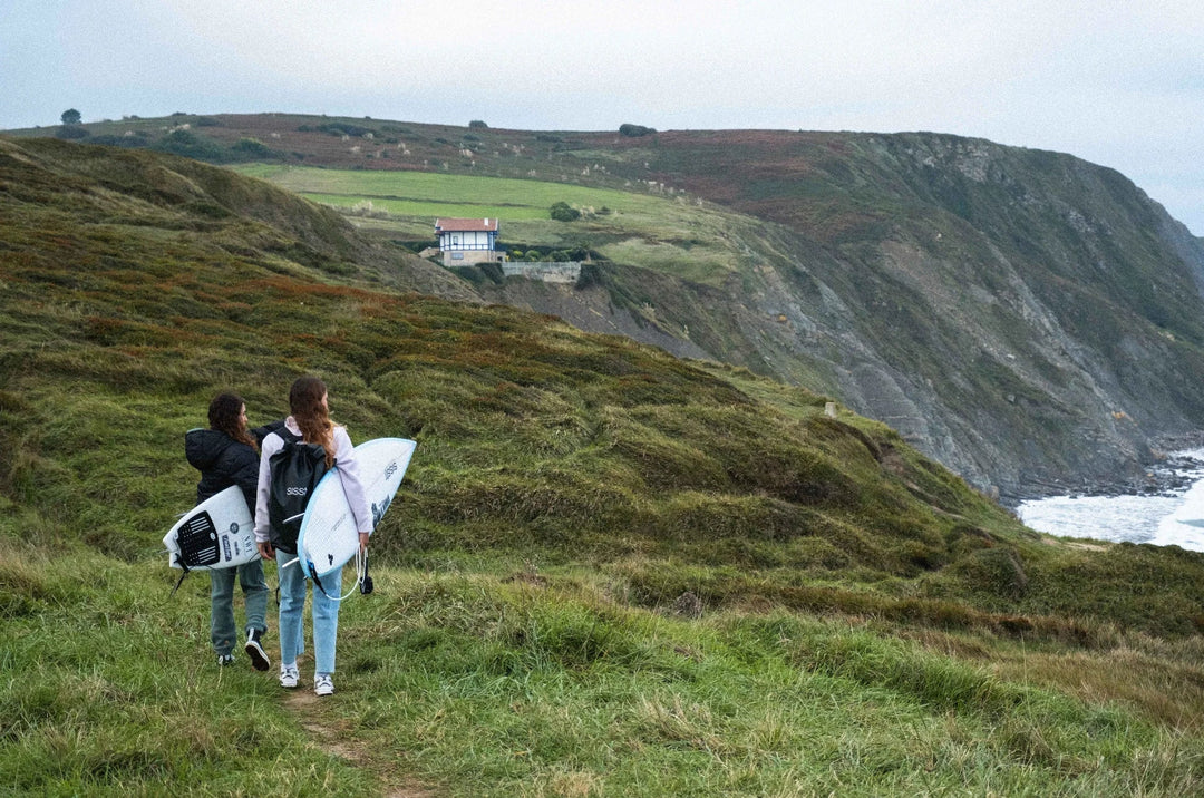 Surfers walking along a grassy cliffside in Northern Spain, overlooking the ocean.