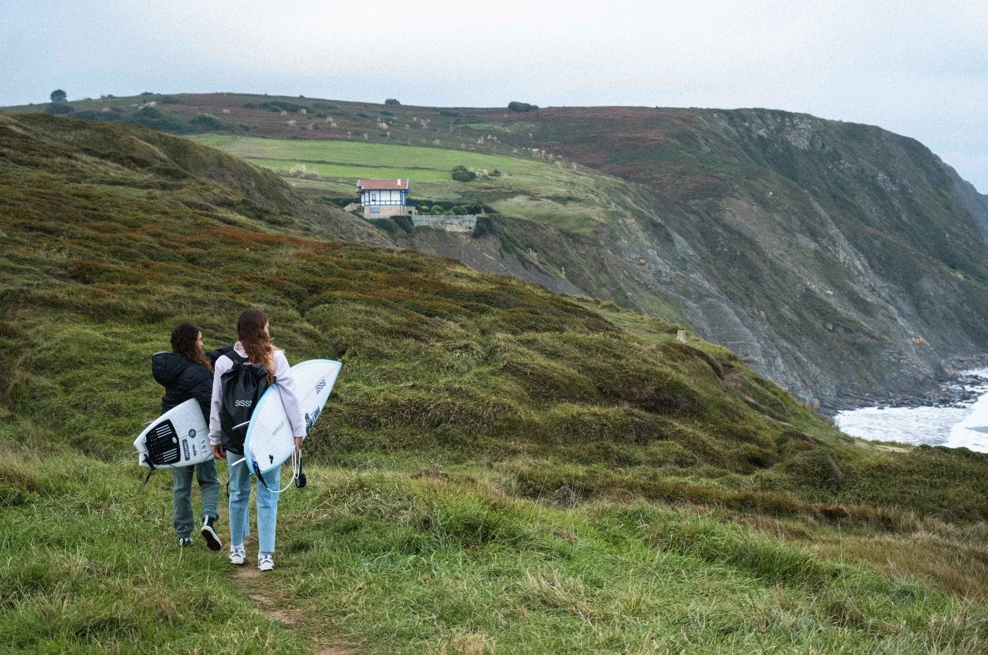 Surfers walking along a grassy cliffside in Northern Spain, overlooking the ocean.