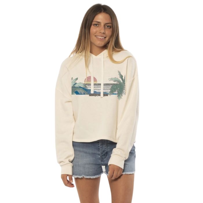 A young woman with long hair is smiling while wearing a Sisstrevolution Sisstr Lines Ls Crop Knt Flc Hoodie, which showcases a sunset panorama with the ocean and palm trees. She is also wearing denim shorts and standing against a plain white background.