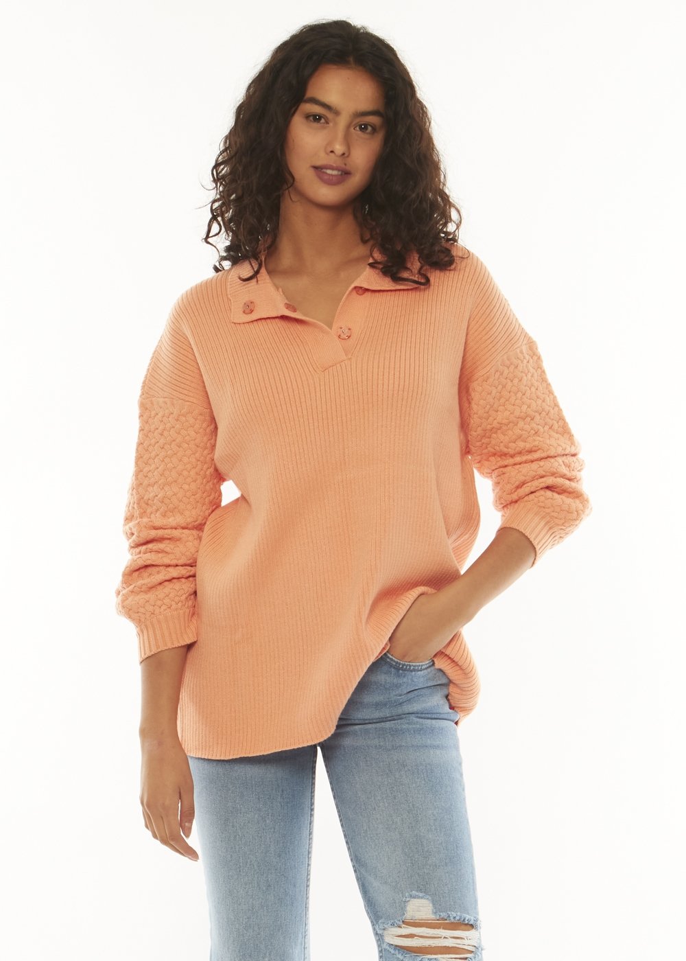 A woman with curly dark hair is wearing a sorbet-colored Sisstrevolution Flint Ls Sweater and ripped blue jeans. She is standing against a plain white background with one hand in her pocket and a relaxed expression.