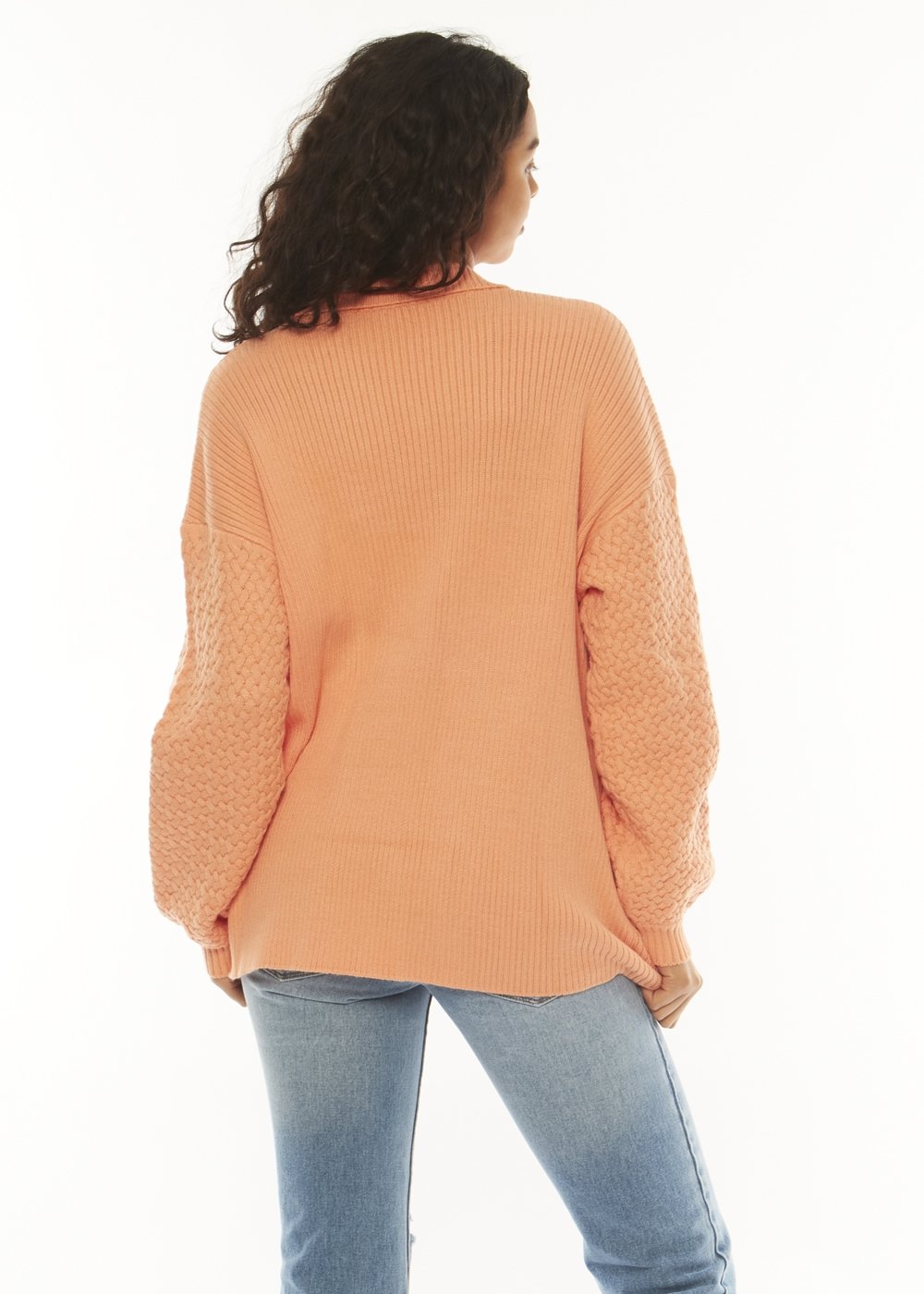 A woman with long curly hair stands with her back to the camera, wearing a sorbet-colored Sisstrevolution Flint Ls Sweater featuring textured sleeves and snap closures, paired with light blue jeans. The background is white and minimalist.