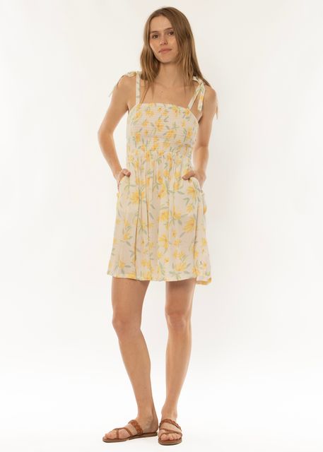 A person with long hair stands against a plain white background, wearing the Sisstrevolution Monte Vista Woven Tank Dress in Sea Salt, which is sleeveless and features a light-colored fabric with a yellow floral pattern. They have their hands in the dress pockets and are wearing brown sandals from Sisstrevolution.