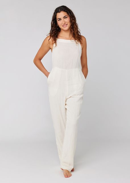 A woman with long, wavy hair is smiling and standing against a plain background. She is wearing the Sisstrevolution Happy Here romper in Sea Salt, a sleeveless organic cotton outfit with adjustable straps and no shoes. With her hands in the pockets, she appears relaxed and comfortable.