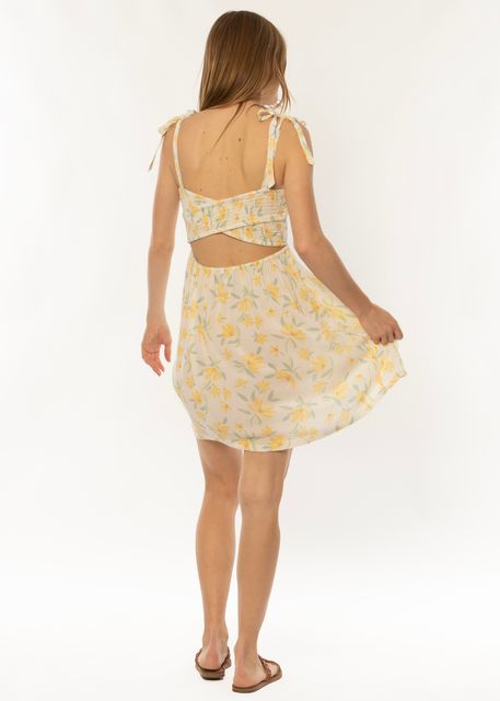 A person with long hair is shown from the back wearing a Sisstrevolution Monte Vista Woven Tank Dress in Sea Salt, featuring tie straps and a cut-out detail at the back. The light, floral dress is above knee-length with a flowy skirt. They are wearing flat sandals and standing against a plain white background.