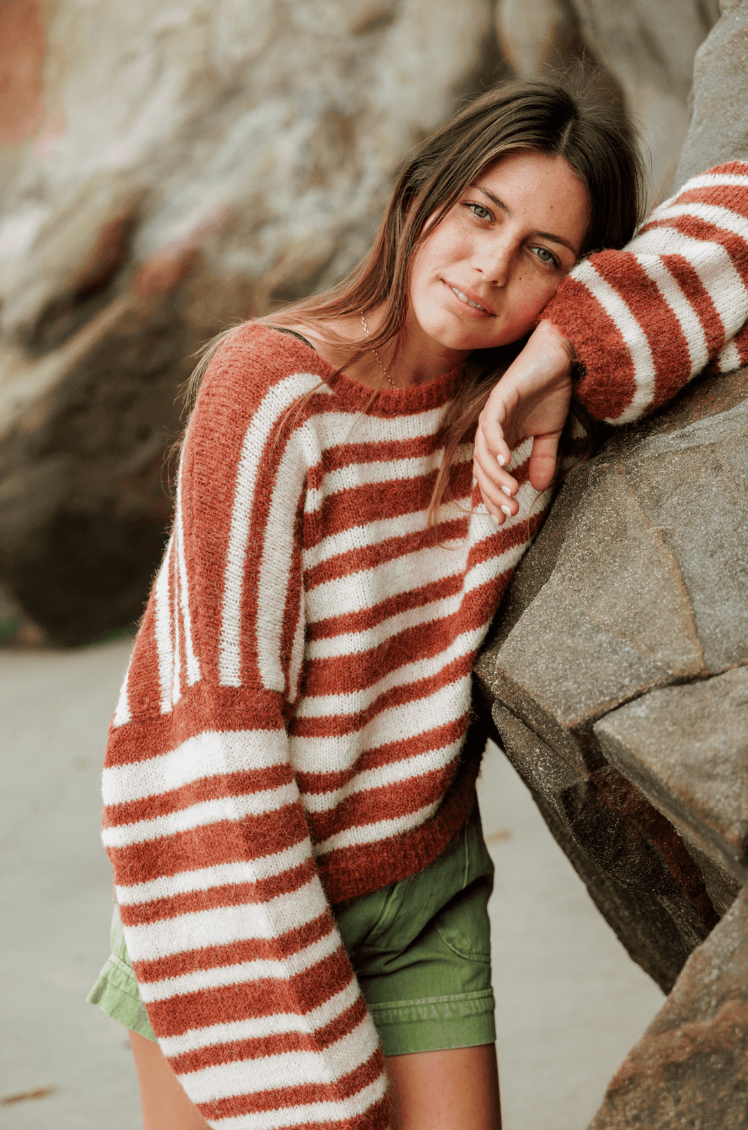Cozy Sisstr Ocean Shores Sweater in rust, paired with green shorts near rocks.
